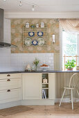 Cream-coloured kitchen cupboards below splashback with subway tiles, floral wallpaper and plate rack