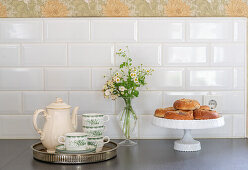 Coffee pot, cups, small bouquet of flowers and rolls on worksurface below splashback with subway tiles