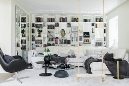 Weißes Wohnzimmer mit Regalwand, Sofagarnitur, schwarzem Tischset, Sessel und Schaukel