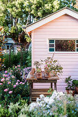 Rustikaler Pflanztisch in blühendem Garten mit rosa Holzhäuschen