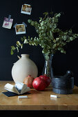 Tisch mit Mörser, Granatäpfeln, Vase und Eukalyptuszweigen vor schwarzer Wand
