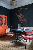 Chairs with pink cover around round table and orange sideboard in room with black wall and stucco