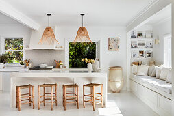 Weiße Küche mit Holzhockern um Kücheninsel, Rattan-Hängeleuchten und eingebauter Sitzbank vor dem Fenster