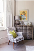 Upholstered armchair in front of window and a vintage chest of drawers in the corner of room