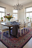 Large dining table with wooden top and white chairs on Persian carpet in bright dining area