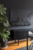 Langes Sideboard, darüber Kunstwerk an schwarzer Wand, im Vordergrund Zimmerpflanze