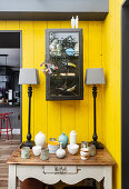 Side table with collection of vases and lamps in front of a yellow-painted wood paneled wall