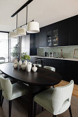 Dunkler, ovaler Esstisch mit Keramiksammlung und Polsterstühle vor Küchenzeile mit schwarzen Schrankfronten