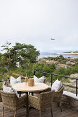 Round table with rattan chairs and cushions on terrace, view of coastal landscape