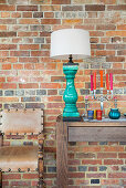 Holzkonsole mit türkisblauer Tischlampe und Kerzenständer neben Lederstuhl vor Backsteinwand