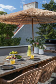 Langer Holztisch mit Obst und Erfrischugnsgetränk, Rattanstühle und Sonnenschirm auf der Dachterrasse