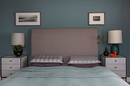 Double bed with high headboard, flanked by bedside tables with lamps in front of grey wall
