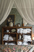 Vintage bedroom with open wooden chest of drawers, patterned wallpaper and fabric canopy