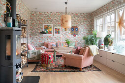 Wood-burning stove and firewood shelf in living room with pink upholstered furniture and vintage wallpaper, low board under the window