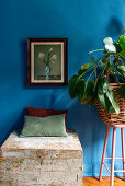 Truhe mit Kissen, Zimmerpflanze und Gemälde an blauer Wand im Schlafzimmer