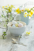 Pastellfarbene Ostereier in Keramikschalen, im Hintergrund Frühlingsstrauß aus Narzissen (Narcissus) und Schlüsselblumen (Primula veris), Obstblütenzweig