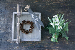 Wooden tray with larch cone wreath and silver fork, bouquet of snowdrops (Galanthus) with ivy leaves and name tag