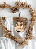 Weiße Kleid und alte Weihnachtskarte auf Kleiderbügel, umrahmt von herzförmigem Kranz mit getrockneten Blüten