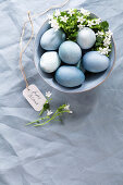 Blue coloured Easter eggs in a ceramic bowl with flower decoration on a blue linen tablecloth