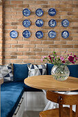 Essbereich mit blauer Eckcouch, Ziertellern an Backsteinwand und Blumenstrauß auf Tisch