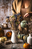 Dried flowers in a white vase on a wooden table with pumpkins and craft materials, autumn decoration