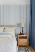 Wooden bed and bedside table in front of blue and white striped wallpaper