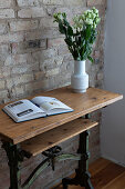 Holztisch mit antikem Untergestell, Buch und Blumenstrauß, Ziegelwand