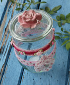 Self-sewn ribbons made of rose fabric, mini roses and fabric rosettes as decoration for storage jar and glass lid with rose on blue wooden table
