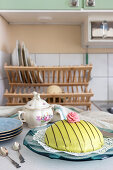 Green princess cake on kitchen table with crockery rack in the background