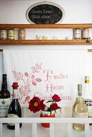Bottles of drinks and flowers in a mug in front of a cloth embroidered with a motto with a wooden spice shelf above