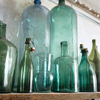 Glass bottles and jars in green on wooden shelf in front of unplastered wall