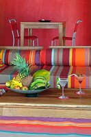 Outdoor furniture upholstered in bright stripes and bowls with tropical fruit on a wooden table; in the background a dining table in front of a bright, red wall