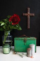 Vintage kitchen utensils in front of green metal case and red flower in vase and cross hanging on black wall