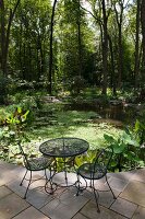 Wire mesh garden table and chairs on stone-flagged terrace with view of pond in woodland garden