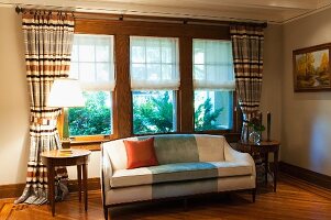 Pale sofa and round side tables in front of window with half-opened blinds and gathered curtains in traditional living room