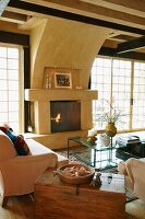 Old wooden trunk between couch and armchair around glass table and open fireplace in living room with lattice terrace doors