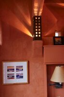 Framed picture below sconce lamp on wall bracket with perforated metal lampshade throwing dramatic pattern of light and shade onto salmon pink wall