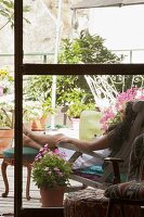 View through French windows onto terrace with greenery - woman sitting on lounger with feet propped up on antique stool