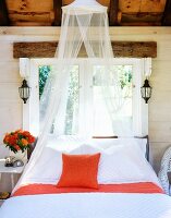 Moskitonetz über französischem Bett am Fenster eines rustikalen Holzhauses