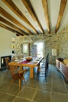 Set dining table with light and dark wooden chairs in Mediterranean dining room with stone walls and wood-beamed ceiling