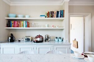 Blick über Marmortischplatte auf eingebautes Sideboard in Wandnische mit zwei weißen Wandboards für Bücher und Wohnaccessoires mit Vintage-Flair