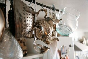 Ornate metal teapot, glass jug and kitchen utensils hanging from rail suspended from ceiling