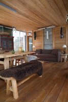 Rustic chalet interior; dining area with dark animal-skin blanket on bench and comfortable sofa next to sliding glass door in background