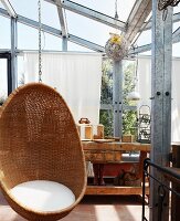 Comfortable, wicker hanging chair with white seat cushions and rustic workbench in conservatory