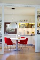 Blick auf weißen runden Tisch mit roten Designerstühlen in weißer Einbauküche mit gelb beleuchteten Hängeschränken