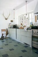 Moderne Küchenzeile vor Fenster, im Hintergrund an Wand aufgehängtes Geweih, in Küche mit ländlichem Flair, Fliesenboden mit einzelnen dunklen Fliesen