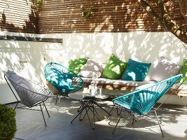 Retro, outdoor cord chairs in various colours on sunny terrace in front of bench with green and blue cushions against half-height wall