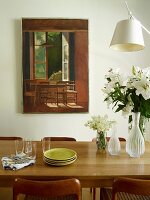 Esstisch aus Eichenholz, darauf Tellerstapel und Glasvasen, mit weißem Lilienstrauss, im Hintergrund an Wand Gemälde