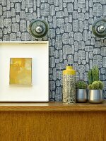 Vase next to cacti and picture on wooden cabinet against wall with retro wallpaper