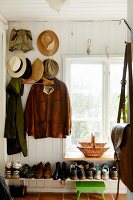 Jackets and hats hung from coat pegs on white wooden wall above full shoe rack below window